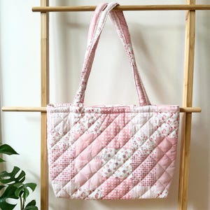 May include: A quilted tote bag featuring a patchwork design in shades of pink and white. The fabric displays a mix of floral patterns, gingham checks, and solid pink squares. It has two matching quilted shoulder straps and a diamond-stitched texture.