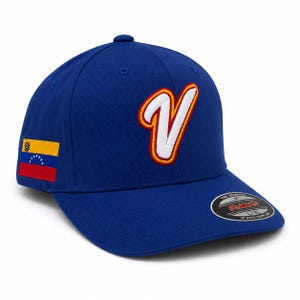 Könnte beinhalten: Eine königsblaue Baseballkappe mit einem großen, weiß gestickten Buchstaben V mit roter und gelber Umrandung auf der Vorderseite. An der Seite befindet sich ein rechteckiger Aufnäher der venezolanischen Flagge und ein schwarzer Aufkleber auf dem Schirm mit der Aufschrift Flexfit.