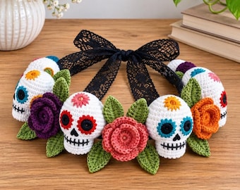 Op de afbeelding: Een gehaakte hoofdband met witte schedels met kleurrijke geborduurde gezichtsdetails, afgewisseld met gehaakte bloemen in paars, roze en oranje, groene bladeren en een zwart kanten lint aan de achterkant voor een goede pasvorm.
