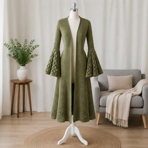 May include: An olive green, long-sleeved, open-front cardigan featuring a crochet knit construction. The garment has dramatic, flared bell sleeves with an intricate diamond-patterned openwork design and a matching flared hemline with a lattice texture.
