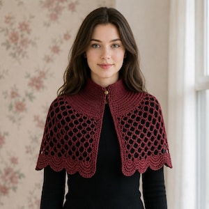 May include: A dark red crochet capelet featuring a solid high collar with a single gold-toned button closure, a decorative openwork mesh pattern across the shoulders and chest, and a scalloped edge finish along the bottom hemline.
