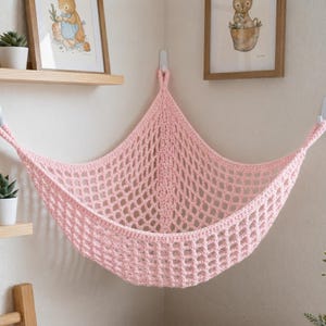 May include: A light pink crochet toy hammock featuring a wide, open-grid mesh pattern. The triangular storage net is designed to be suspended in a corner, with three reinforced loops at the corners for mounting to wall hooks or hardware.