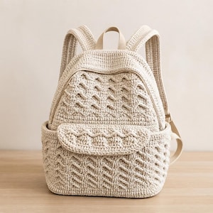 Puede incluir: Una mochila de ganchillo color crema con un patrón de ondas texturizado en el cuerpo principal y el bolsillo delantero. La bolsa incluye un asa superior, dos correas ajustables para los hombros y un cierre de cremallera, todo en hilo neutro.