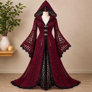 May include: A floor-length, deep burgundy crochet hooded coat featuring wide bell sleeves and a black scalloped trim along the edges. The garment is secured at the front with three ornate, metallic-toned filigree clasps, creating a structured silhouette.