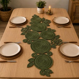 Peut inclure: Un chemin de table en crochet vert olive foncé composé d'une série de napperons circulaires reliés par des motifs floraux et des feuilles. Le chemin est centré sur une table en bois clair avec quatre assiettes blanches et des couverts.