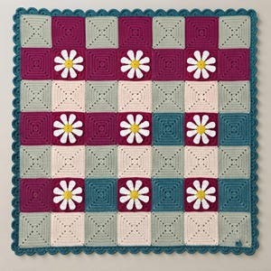 May include: A square crochet blanket featuring a grid of alternating magenta, teal, and cream-colored squares. Several squares are adorned with white and yellow daisy appliques. The entire blanket is finished with a scalloped teal border.