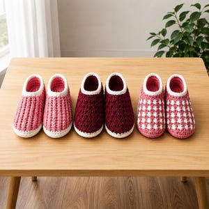 May include: Three pairs of handmade crochet slippers in different colours and textures. The first pair is light pink with vertical ridges, the second is deep burgundy with a textured stitch, and the third is a pink and white diamond pattern.
