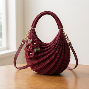 May include: A deep burgundy crochet handbag with a crescent shape and ribbed texture. The bag features a top handle, a detachable shoulder strap with gold-tone hardware, and a cluster of three dark red crocheted flowers with green leaves.