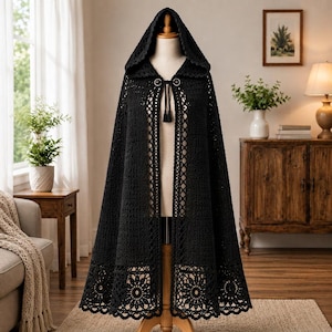 Op de afbeelding: Een zwarte gehaakte cape met capuchon, voorzien van een ingewikkeld opengewerkt patroon langs de voorranden en een gedetailleerde bloemenrand aan de zoom. Het kledingstuk wordt bij de hals gesloten met een decoratieve sluiting en twee kwastjes.