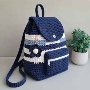 Pode incluir: Uma mochila de crochê às riscas azul-marinho e branca com alça superior, alças de ombro ajustáveis e um bolso frontal com fecho branco. Um pendente em forma de flor azul-escura e branca está preso a um fecho dourado na aba frontal.