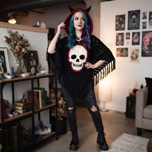 以下が含まれることがあります： 胸元に大きなクリーム色のスカルのアップリケ、フードに小さな黒い角、裾に長いフリンジがあしらわれた、黒いかぎ針編みのフード付きポンチョ。ダメージ加工の黒いデニムジーンズと黒いブーツを合わせて着用。