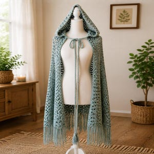 May include: A sage green crochet hooded cape featuring an openwork stitch pattern, a pointed hood, and long, thin braided ties at the neck. The garment is finished with a straight hemline adorned with long, dangling fringe along the bottom edge.