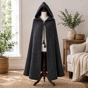 May include: A dark charcoal gray hooded cloak featuring a thick, textured crochet knit pattern. The garment includes a single wooden button closure at the neck with two long, braided cord ties that hang down, each finished with a small tassel at the end.