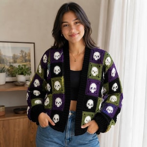 May include: A long-sleeve crochet cardigan featuring a checkerboard pattern of black, purple, and olive green squares. Each square contains a white skull motif. The garment has a solid black ribbed trim along the front opening and cuffs.