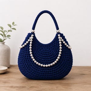 Può includere: Una borsa all'uncinetto blu navy scuro con una silhouette arrotondata e un manico intrecciato singolo. La borsa è decorata con due fili drappeggiati di perle sferiche color argento lucido fissati alla base del manico.