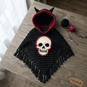 Puede incluir: Un poncho de ganchillo negro con capucha, dos cuernos pequeños, forro interior rojo y una gran aplicación de calavera color crema con borde rojo en la parte delantera. La prenda tiene flecos a lo largo del borde inferior.