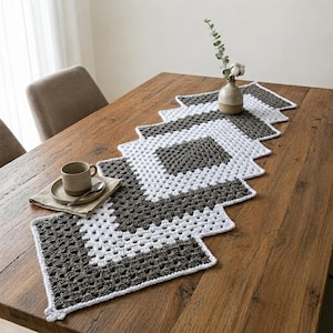 Puede incluir: Un camino de mesa de ganchillo rectangular con un patrón repetitivo de cuadrados granny en hilo gris oscuro y blanco alternados. El camino está extendido, con un jarrón de cerámica con una rama de eucalipto y una taza de cerámica en un plato.