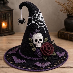 Könnte beinhalten: Ein schwarzer, gehäkelter Spitzhut mit weißem Spinnennetzmuster, einem weißen Totenkopf, einer dunkelroten Rose, grünen Blättern und violetten Fledermäusen am Rand. Eine kleine schwarze Spinne hängt an einem weißen Faden an der Spitze.