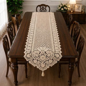 May include: A long, rectangular cream-coloured lace table runner featuring an intricate floral pattern and a geometric mesh centre. The runner has scalloped edges and a single decorative tassel hanging from the pointed end, centred on a dark wood table.