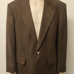 May include: A brown blazer featuring a small-scale houndstooth pattern throughout. The garment includes a notched lapel, a single-button front closure, two flap pockets at the waist, and a single welt pocket on the upper left chest area.