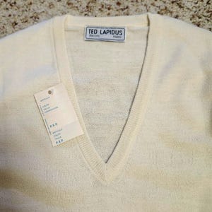 May include: A cream-colored V-neck sweater featuring a ribbed neckline and a white brand label at the collar that reads TED LAPIDUS TRICOTS PARIS. A rectangular paper hang tag is attached to the front, displaying printed text in blue ink.