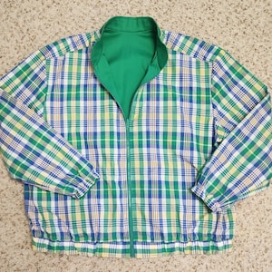 May include: A reversible zip-up jacket featuring a plaid pattern in shades of green, blue, white, and yellow on one side. The jacket has a high collar, long sleeves with elastic cuffs, and a solid green interior lining visible at the neckline.