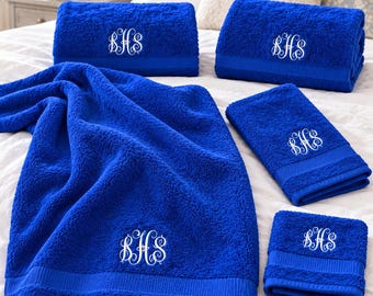 May include: A set of four royal blue terry cloth towels of varying sizes, each featuring a white embroidered monogram with the letters BHS in a decorative script font. The towels have a textured surface and a smooth horizontal band detail.