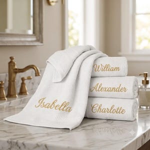 May include: A stack of three white cotton towels with gold embroidered names, including William, Alexander, and Charlotte, plus a draped white towel with the name Isabella embroidered in gold script, all featuring a soft, textured fabric finish.