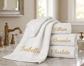May include: A stack of three white cotton towels with gold embroidered names, including William, Alexander, and Charlotte, plus a draped white towel with the name Isabella embroidered in gold script, all featuring a soft, textured fabric finish.