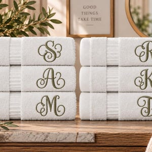May include: A set of six white cotton bath towels stacked in two columns of three. Each towel features a textured border and is embroidered with a large, ornate olive green letter in a cursive script, displaying the letters S, A, M, R, K, and T.