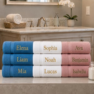 May include: A stack of nine rectangular bath towels arranged in three columns and three rows. The towels are colored blue, white, and dusty rose, each featuring a name embroidered in gold thread: Elena, Sophia, Ava, Liam, Noah, Benjamin, Mia, Lucas, and Isabella.