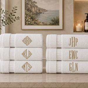 May include: A set of six white cotton towels stacked in two columns. The left stack features three towels with gold embroidered diamond-shaped monograms. The right stack features three towels with gold embroidered vertical letter monograms.