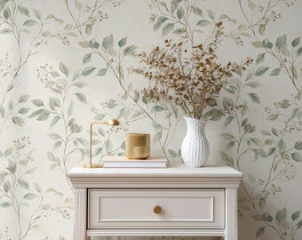 May include: A light-colored wooden nightstand with a single drawer and a gold knob, topped with a white vase containing dried brown branches, a gold candle holder, and a book, set against wallpaper featuring a green leaf pattern.
