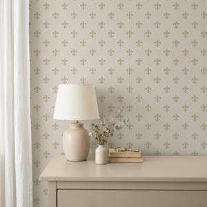 Könnte beinhalten: Eine beige Wand mit einer Tapete, die ein sich wiederholendes goldenes Fleur-de-Lis-Muster aufweist. Vor der Wand steht ein beiger Tisch mit einer beigen Tischlampe mit weißem Schirm, einer kleinen weißen Vase mit Trockenblumen und zwei Büchern.