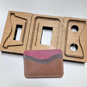 May include: A leather card holder in brown, grey, and pink, with white stitching, next to a wooden die-cut mold. The mold is for crafting leather goods and is made of light brown wood. The card holder is approximately 10 cm long.