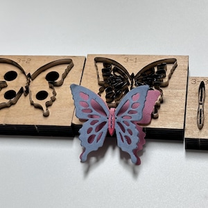 Pode incluir: Uma coleção de peças de artesanato em madeira em forma de borboleta. O conjunto inclui três recortes de borboleta em madeira e uma borboleta azul e rosa em camadas. As peças de madeira estão rotuladas 3-2, 3-1 e 3-3.
