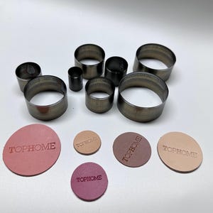 Puede incluir: Un conjunto de cortadores circulares de metal de varios tamaños, junto con varias piezas de cuero circulares. Las piezas de cuero son en tonos de rosa, beige y morado, cada una estampada con la palabra "TOPHOME".