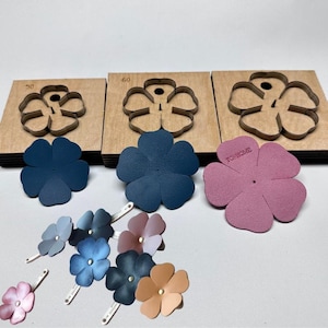 Op de afbeelding: Een verzameling bloemvormige leren uitsparingen in verschillende tinten blauw, roze en bruin. De uitsparingen worden getoond met houten bloemvormige stansmessen. Sommige bloemen hebben metalen haarclips.
