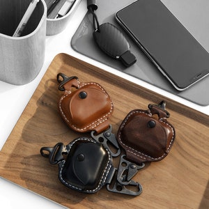 Op de afbeelding: Drie leren Airpod-hoesjes met een karabijnhaak. De hoesjes zijn bruin, zwart en donkerbruin. De hoesjes liggen op een houten dienblad. De tekst "TOPHOME Simple Design Simple Life" staat op het dienblad.
