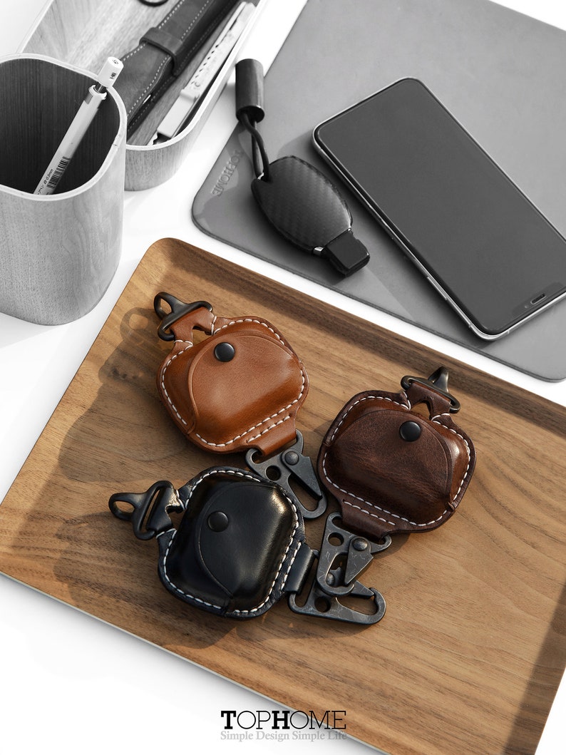 Op de afbeelding: Drie leren Airpod-hoesjes met een karabijnhaak. De hoesjes zijn bruin, zwart en donkerbruin. De hoesjes liggen op een houten dienblad. De tekst "TOPHOME Simple Design Simple Life" staat op het dienblad.