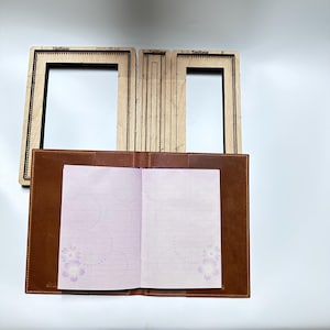 Könnte beinhalten: Ein brauner Leder-Notizbuchumschlag mit einem offenen Heft mit hellvioletten, linierten Seiten, die schwache Kreis- und Blumenmuster aufweisen, platziert unter drei rechteckigen Holzschablonen mit der Aufschrift TenHome und kleinen Metallstiften.
