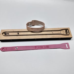 Może przedstawiać: Jasnobrązowa skórzana bransoletka i różowy skórzany pasek, oba z wytłoczonym napisem "TOPHOME". Obecne jest również drewniane narzędzie do wykrawania, przeznaczone do tworzenia tych skórzanych akcesoriów.