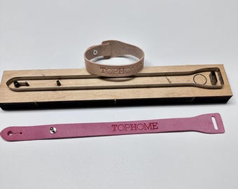 May include: A light brown leather bracelet and a pink leather strap, both with the text "TOPHOME" embossed. A wooden die-cut tool is also present, designed for crafting these leather accessories.