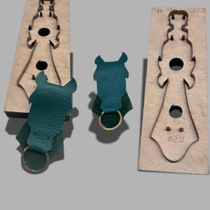 May include: Green leather horse head keychains with metal key rings. The keychains are next to wooden molds with metal outlines of the horse head shape. The molds have the numbers "2.0" and "5.07" printed on them.