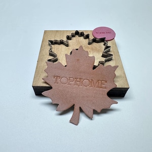Könnte beinhalten: Ein Ahornblatt-förmiger Lederausschnitt mit dem geprägten Text TOPHOME, der auf einem Holzstanzwerkzeug mit passender Metallklingenform liegt, sowie ein kleines rosa kreisförmiges Etikett, das ebenfalls den geprägten Text TOPHOME trägt.