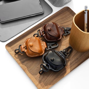 Op de afbeelding: Drie leren hoesjes voor draadloze oordopjes met een karabijnhaak. De hoesjes zijn bruin, beige en zwart. De hoesjes liggen op een houten dienblad met een houten beker en een potlood op de achtergrond.