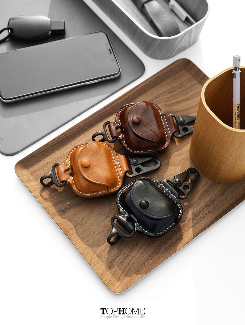 Op de afbeelding: Drie leren hoesjes voor draadloze oordopjes met een karabijnhaak. De hoesjes zijn bruin, beige en zwart. De hoesjes liggen op een houten dienblad met een houten beker en een potlood op de achtergrond.