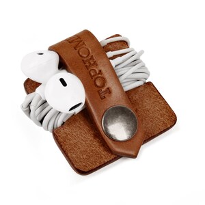 May include: A brown leather cord keeper with a metal button and the word "TOPHON" embossed on the strap. White earbuds are wrapped around the cord keeper.