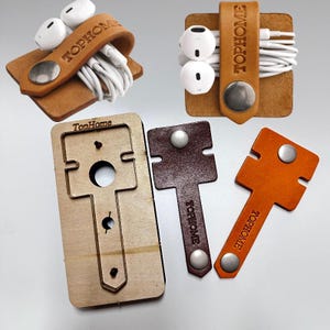 May include: A collection of leather cord organizers featuring the embossed text TOPHOME. The set includes two tan and dark brown leather wraps with silver metal snaps, white wired earbuds, and a wooden cutting die template for crafting.