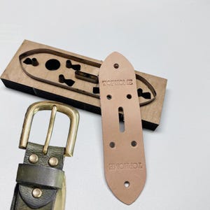 Może przedstawiać: Zielony skórzany pasek z mosiężną klamrą i jasnobrązowym skórzanym paskiem. Na zdjęciu znajduje się również drewniane narzędzie do wykrawania i jasnobrązowy kawałek skóry z napisem "TOPHOME". Klamra jest złota.